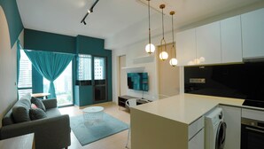 Ein modernes Wohnzimmer mit einer Couch, einem Couchtisch und einem Fernseher. Es gibt eine Küchenzeile mit Kühlschrank, Herd und Mikrowelle. Der Raum hat türkisfarbene Wände und ein großes Fenster mit Vorhängen.