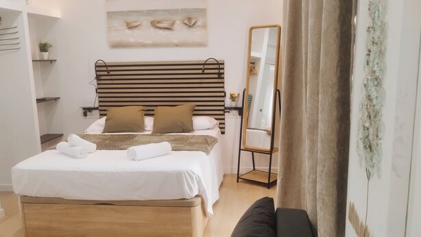 A modern bedroom with a wooden bed, a black sofa, a bedside table, a mirror, and a wall-mounted shelf with a plant. - GO&REST APARTMENTS (València)