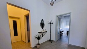 Een hal met een potplant, een wandplank met decoratieve objecten, een vloerlamp en een piano op de achtergrond.