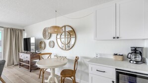 A modern kitchen with white cabinets, a dining table with chairs, a coffee maker, and a TV on a wooden stand. - Ocean View Escape in this 2 bedroom 2 bathroom Condo unit 5 (New Smyrna Beach)