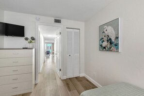 Una habitación con una cama, televisión, tocador blanco, armario y un cuadro en la pared.