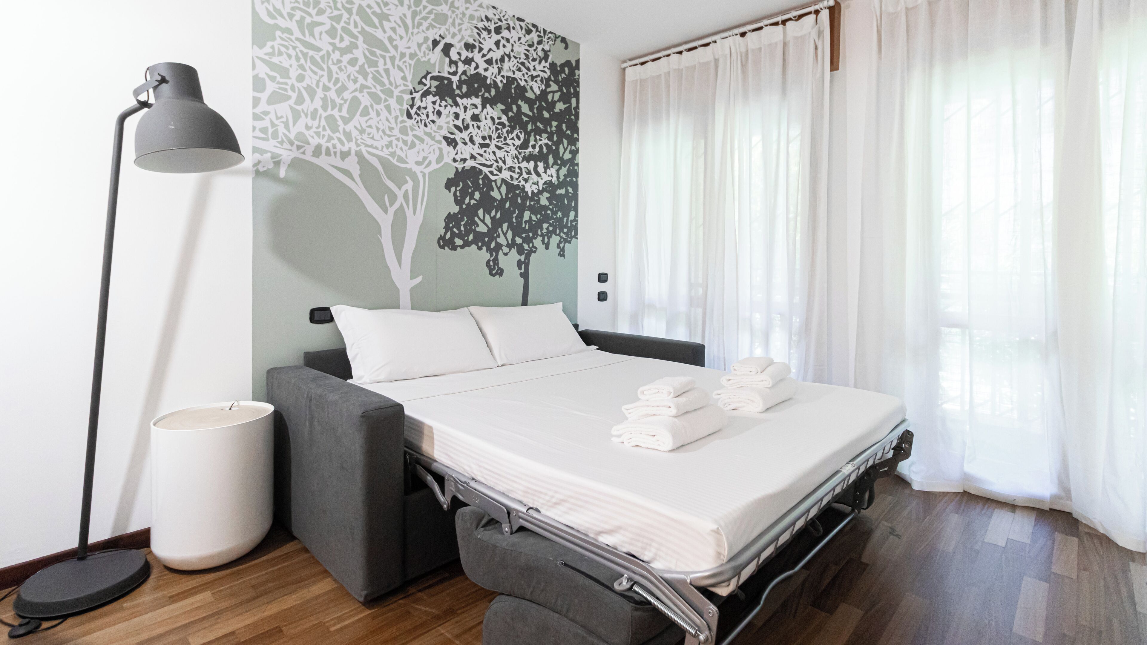 A neatly made bed with white linens and folded towels, a grey sofa, a floor lamp, and a wall mural of a tree.