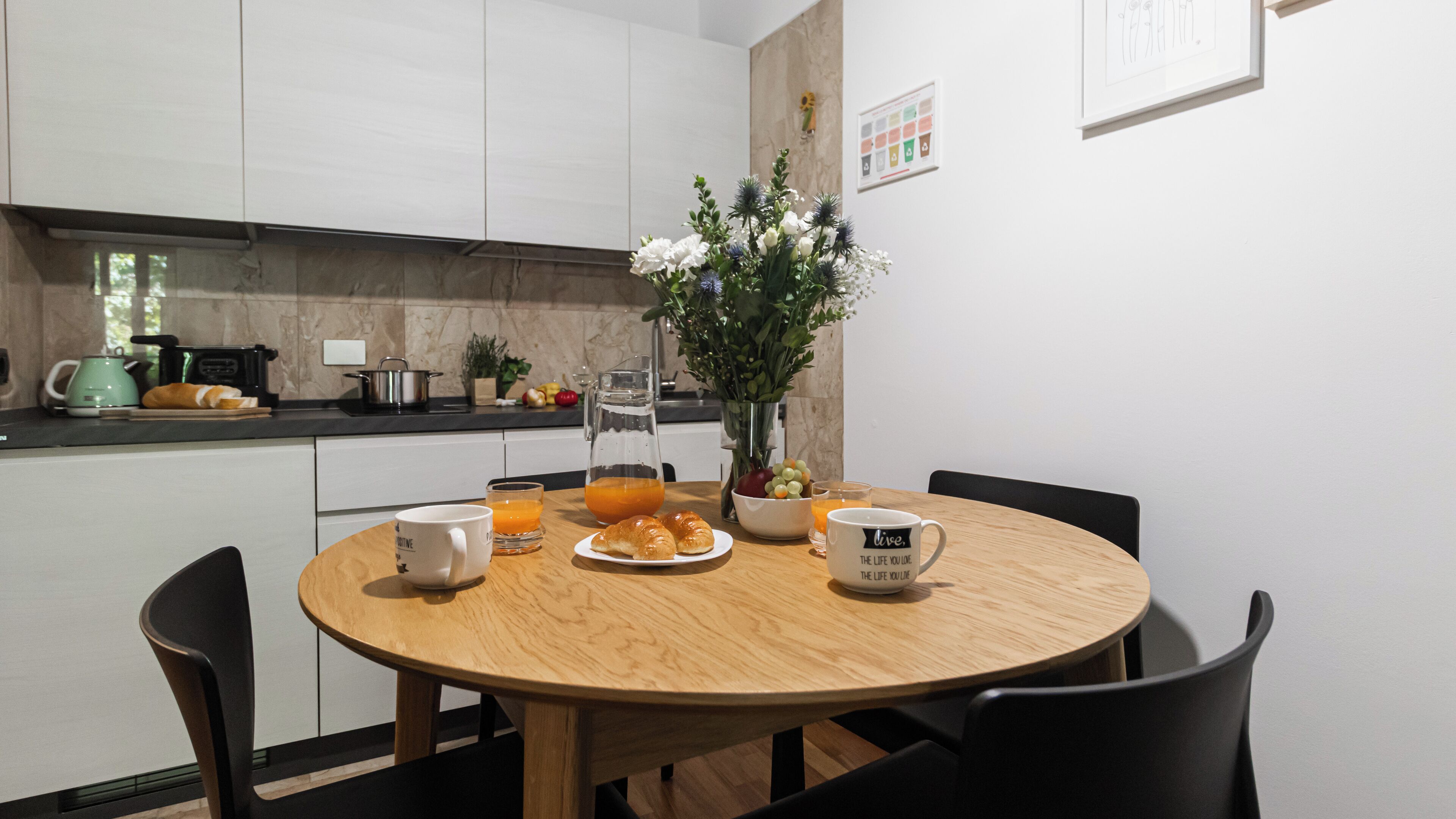 A modern kitchen with white cabinets, a round wooden table, and a vase with flowers.