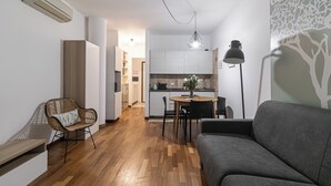 A modern living room with a grey sofa, a wicker chair, and a dining table with chairs. There is a kitchen in the background with white cabinets and a hanging light fixture. - Italianway Comfort - Londonio 23 (Milano)