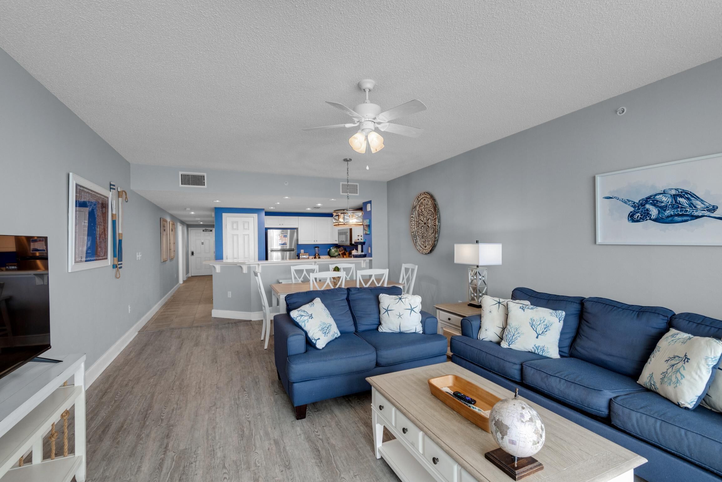 A modern living room with a blue sofa, a coffee table, and a TV. There is a kitchen in the background with white cabinets and a dining area.