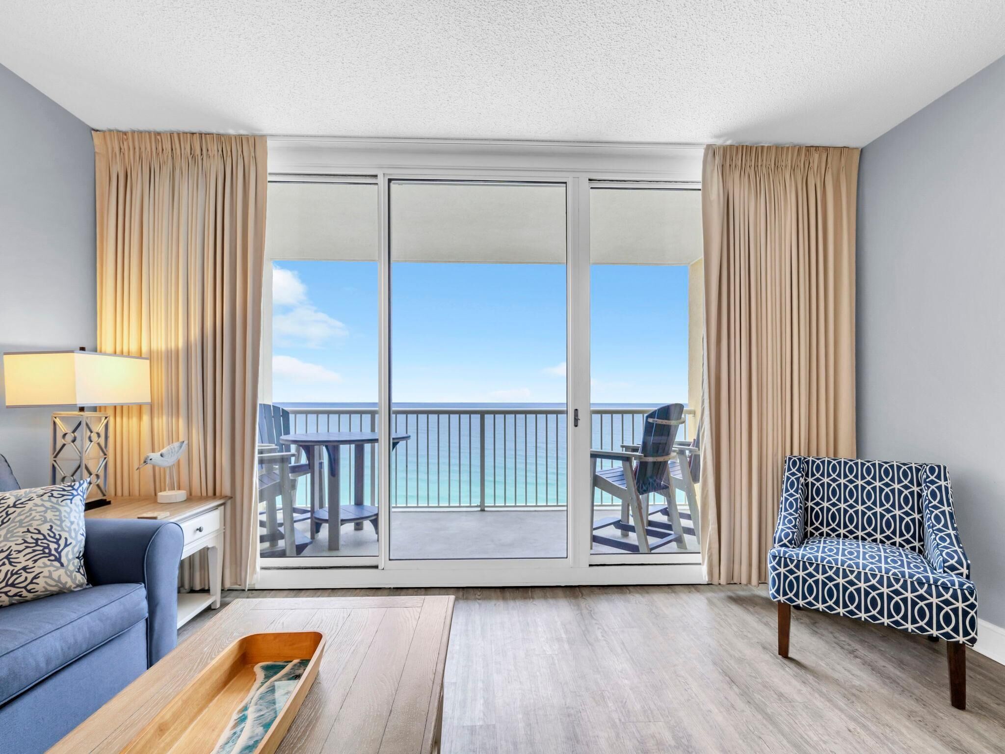 A hotel room with a balcony, a sofa, a patterned armchair, a small table, and a lamp.