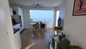 A modern apartment with a kitchen, dining area, and a large window overlooking the sea.