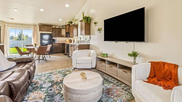 Ein modernes Wohnzimmer mit einer Ledersofa, einem runden Couchtisch und einem weißen Sessel. An der Wand ist ein Flachbildfernseher befestigt, und im Hintergrund befindet sich eine Essecke mit Stühlen sowie eine Küche.