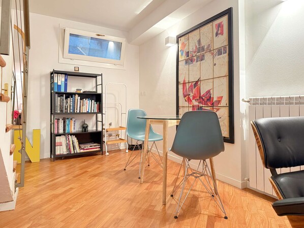 Une pièce avec un plancher en bois, une bibliothèque, une table avec des chaises, un radiateur et un grand tableau abstrait.