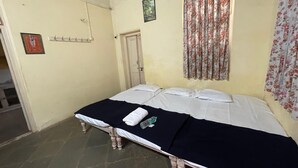 A single bed with a black bedspread and white pillows in a room with cream walls, a door, and a floral curtain. - The Imperial Heritage (Lonavala)