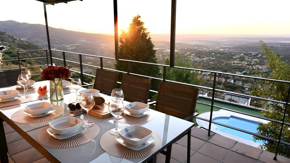 A dining table set with plates, glasses, and a vase of flowers, overlooking a scenic view with a pool.