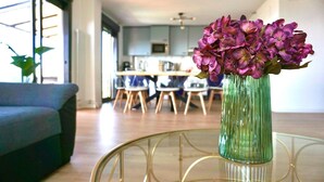 Ein Wohnzimmer mit einer Couch, einem runden Glastisch mit einer Vase voller lila Blumen und einer Küche im Hintergrund.