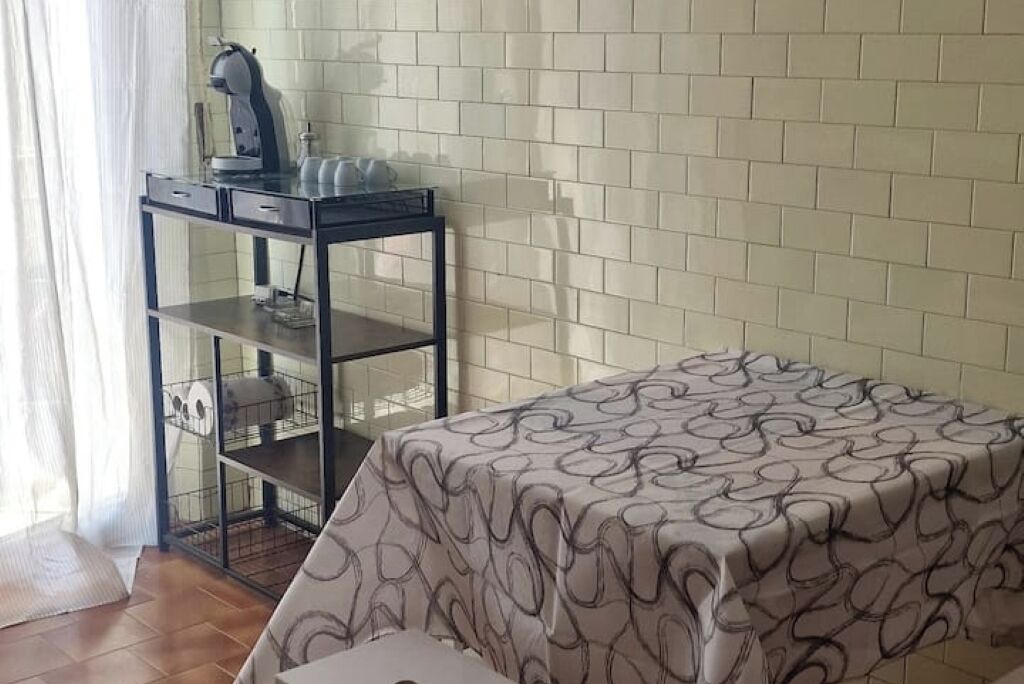 Una stanza con un letto ricoperto da una coperta a motivi decorativi, una scrivania con un bollitore e una scaffalatura con una gabbietta.