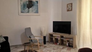 Een woonkamer met een flatscreen televisie, een grijze fauteuil, een zwarte bank en een klein houten tv-meubel met boeken.