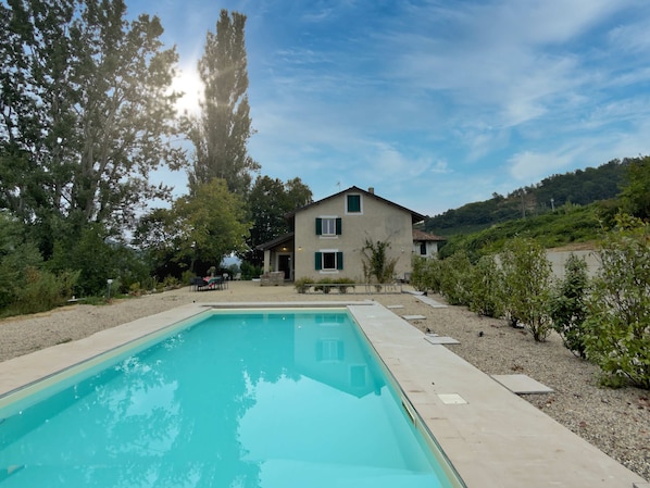 Une maison à deux étages avec une piscine, entourée d’arbres et située dans un paysage vallonné.