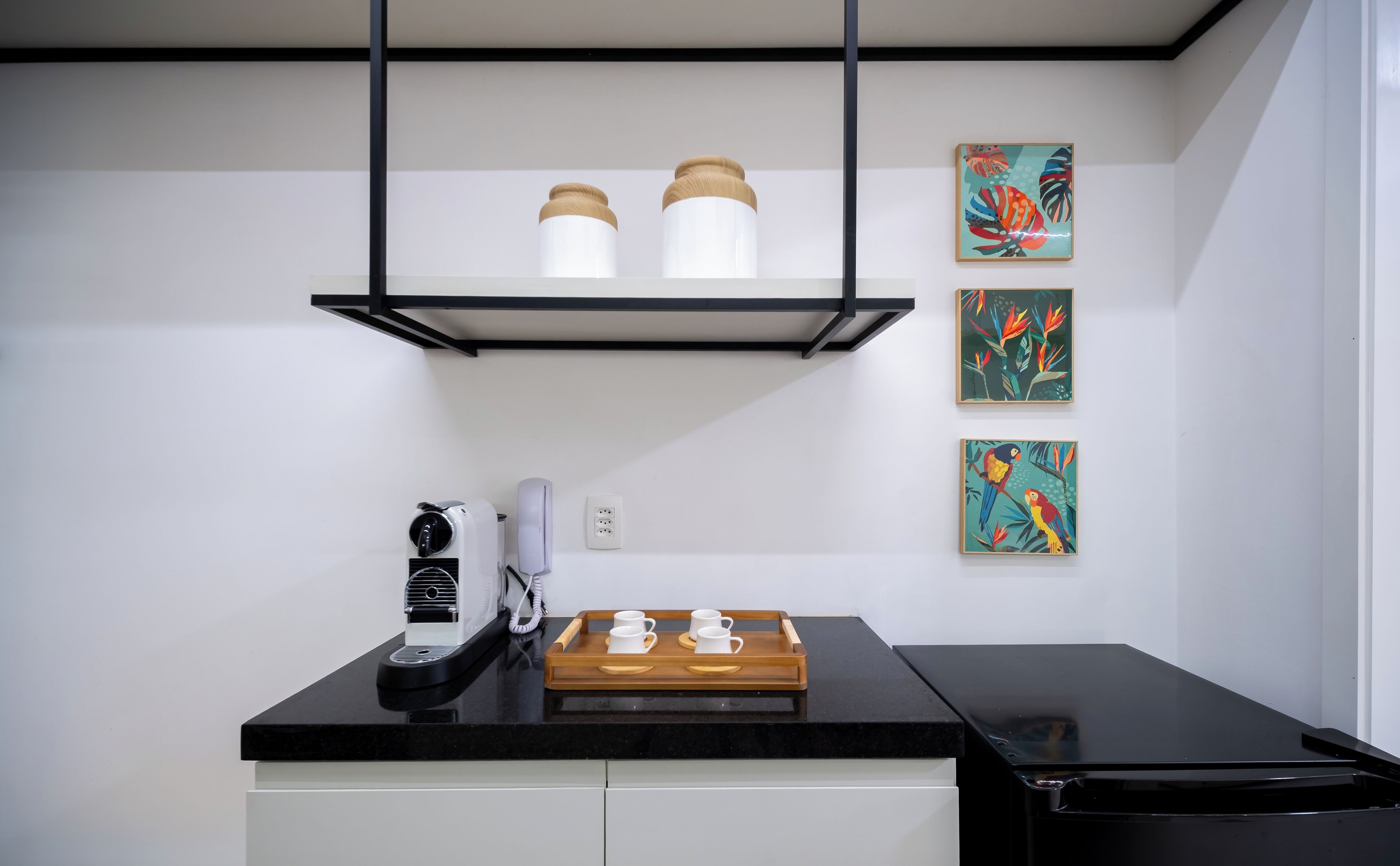 A modern kitchen with a black countertop, a coffee machine, a toaster, and a tray with four cups. There are three wall-mounted shelves above the counter, each holding a white container with a wooden lid.