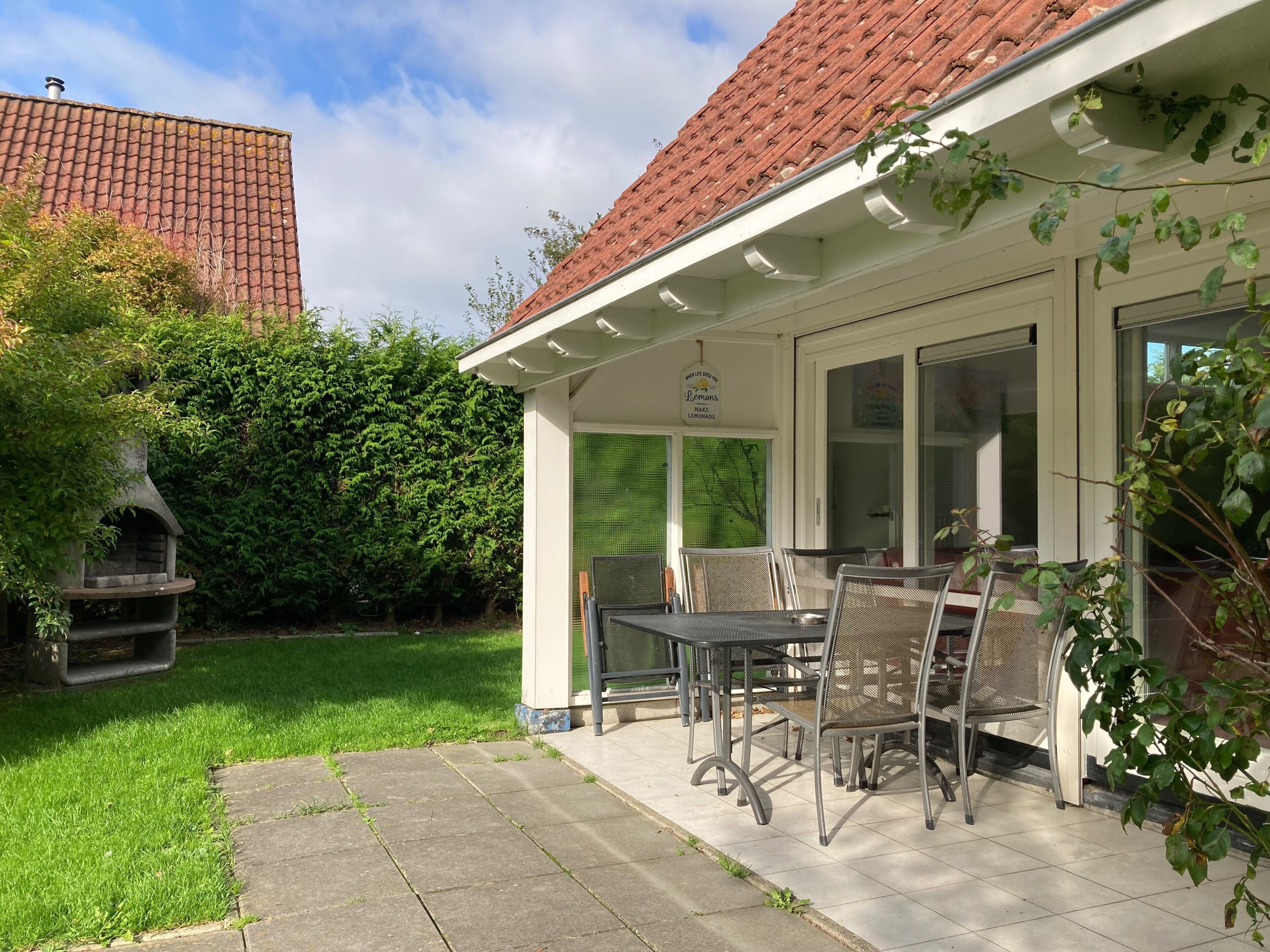A covered patio with outdoor seating, a grill, and a view of a red-tiled roof building.