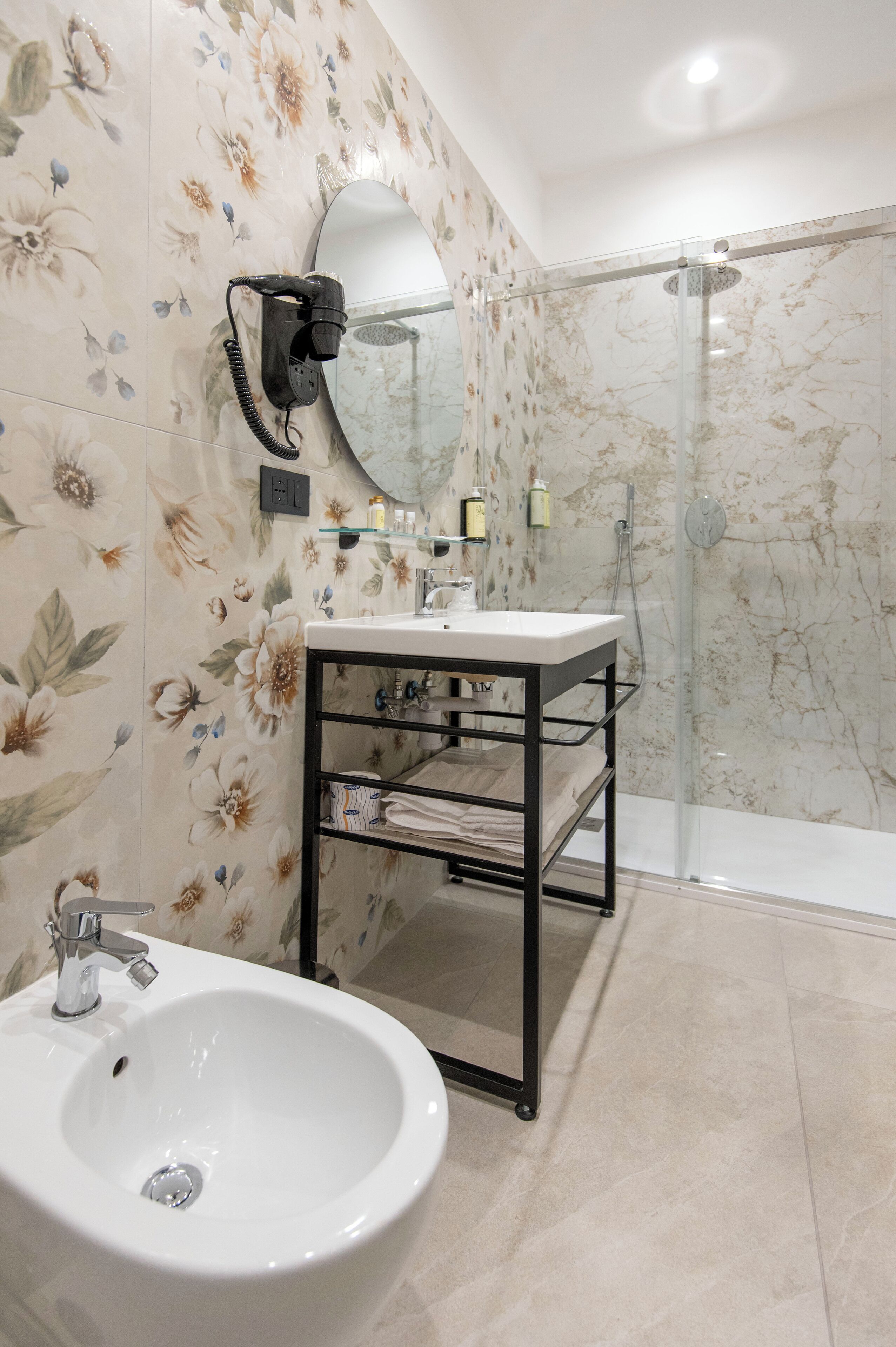 Un baño con un lavabo blanco, un espejo, una mampara de vidrio para la ducha y un papel tapiz con estampado floral.