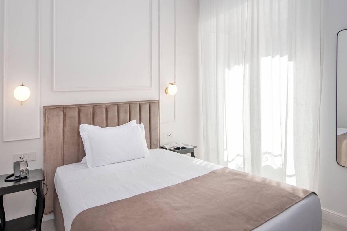 A hotel room with a bed, a bedside table with a phone, a nightstand, and a window with sheer curtains.