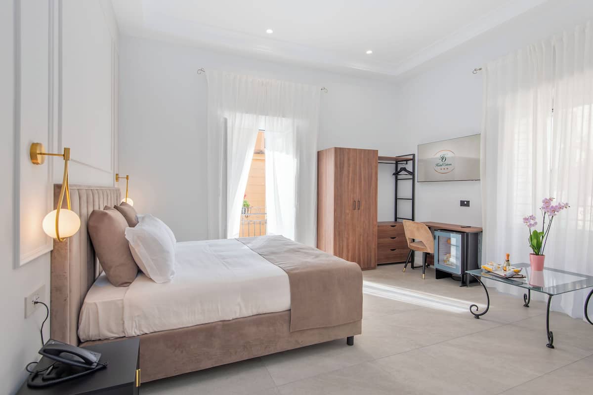 A modern hotel room with a bed, bedside table, lamp, wardrobe, desk, chair, and a small TV.