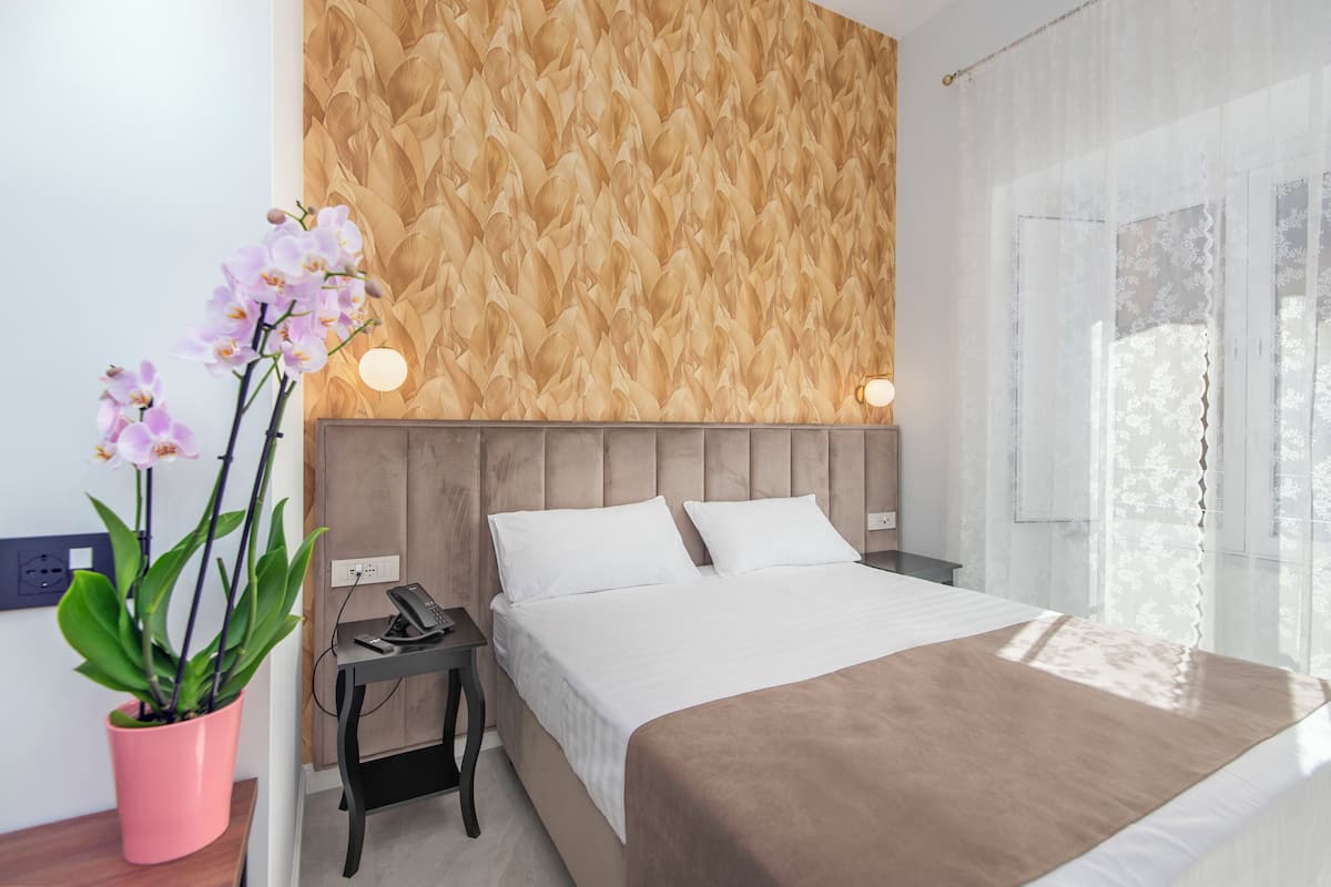 A modern hotel room with a wooden textured wall, a large bed with white linens, a pink orchid plant, and a nightstand with a phone and remote control.