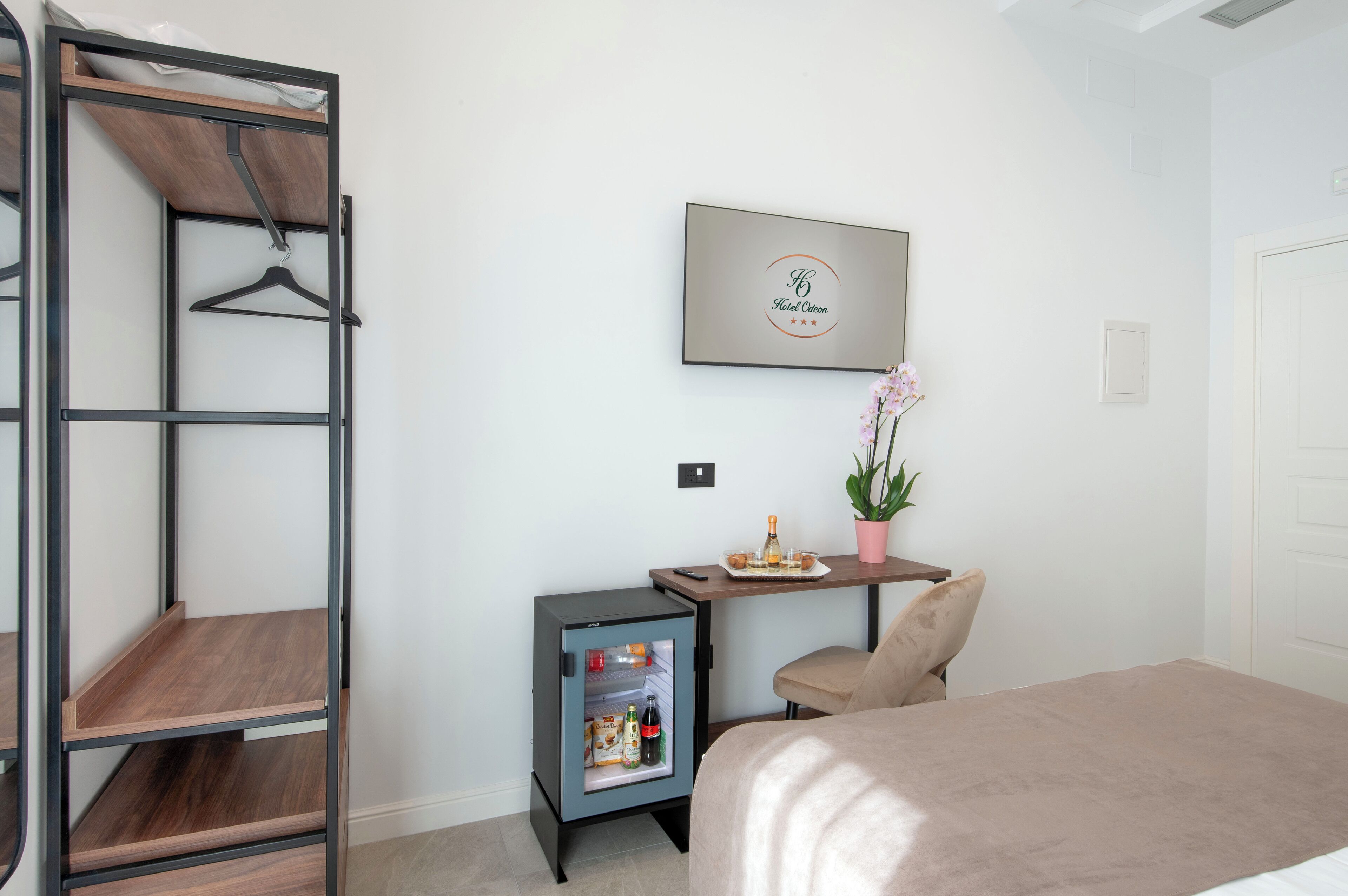 A hotel room with a bed, a small table with a plant, a minibar, and a wall-mounted picture.