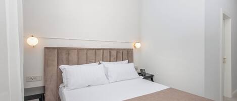 Una cama bien hecha con sábanas blancas y un plaid marrón, una mesita de noche con teléfono y lámpara, y un diseño de habitación sencillo y limpio.