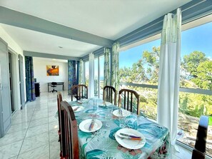 A dining area with a table set for four, large windows, and a view of greenery. - Villa Copernic, Pool and Ocean View (Sainte-Marie)
