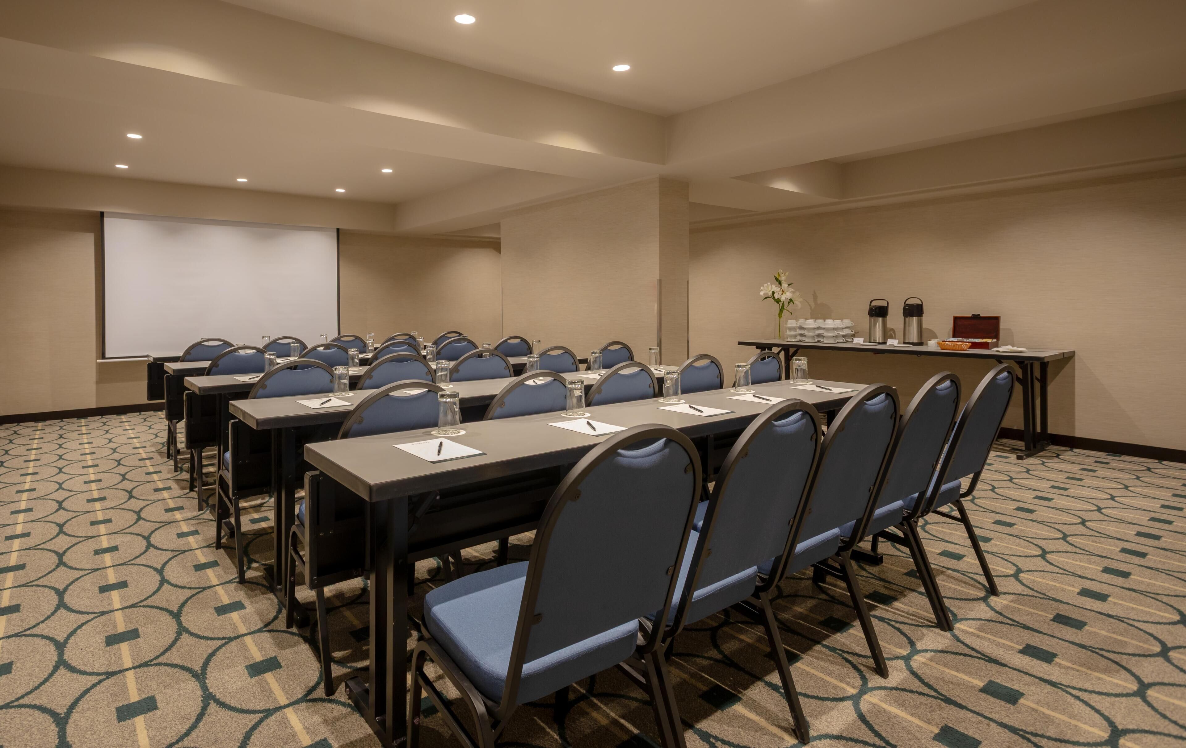 Un salón de conferencias con varias mesas y sillas, una pantalla de proyector y una pequeña mesa con botellas de agua y un jarrón de flores.