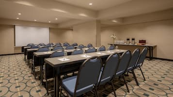 Un salón de conferencias con varias mesas y sillas, una pantalla de proyector y una pequeña mesa con botellas de agua y un jarrón de flores.
