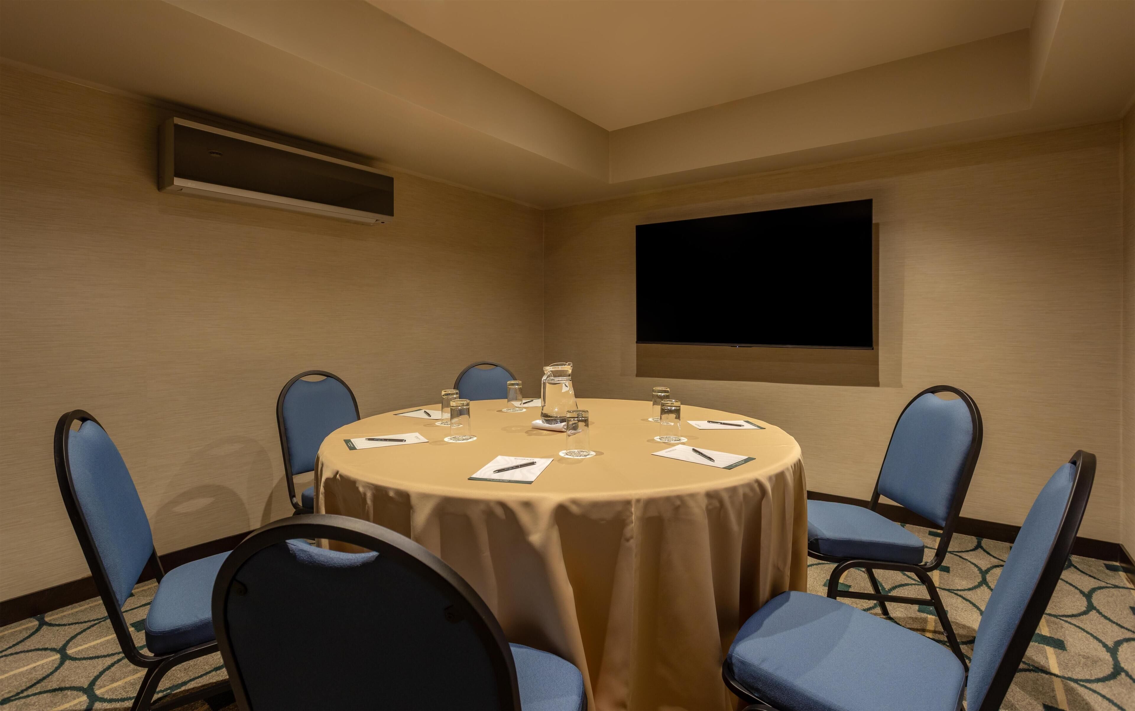 Un salón de conferencias con una mesa redonda, sillas azules, una pantalla de proyector y una jofaina.