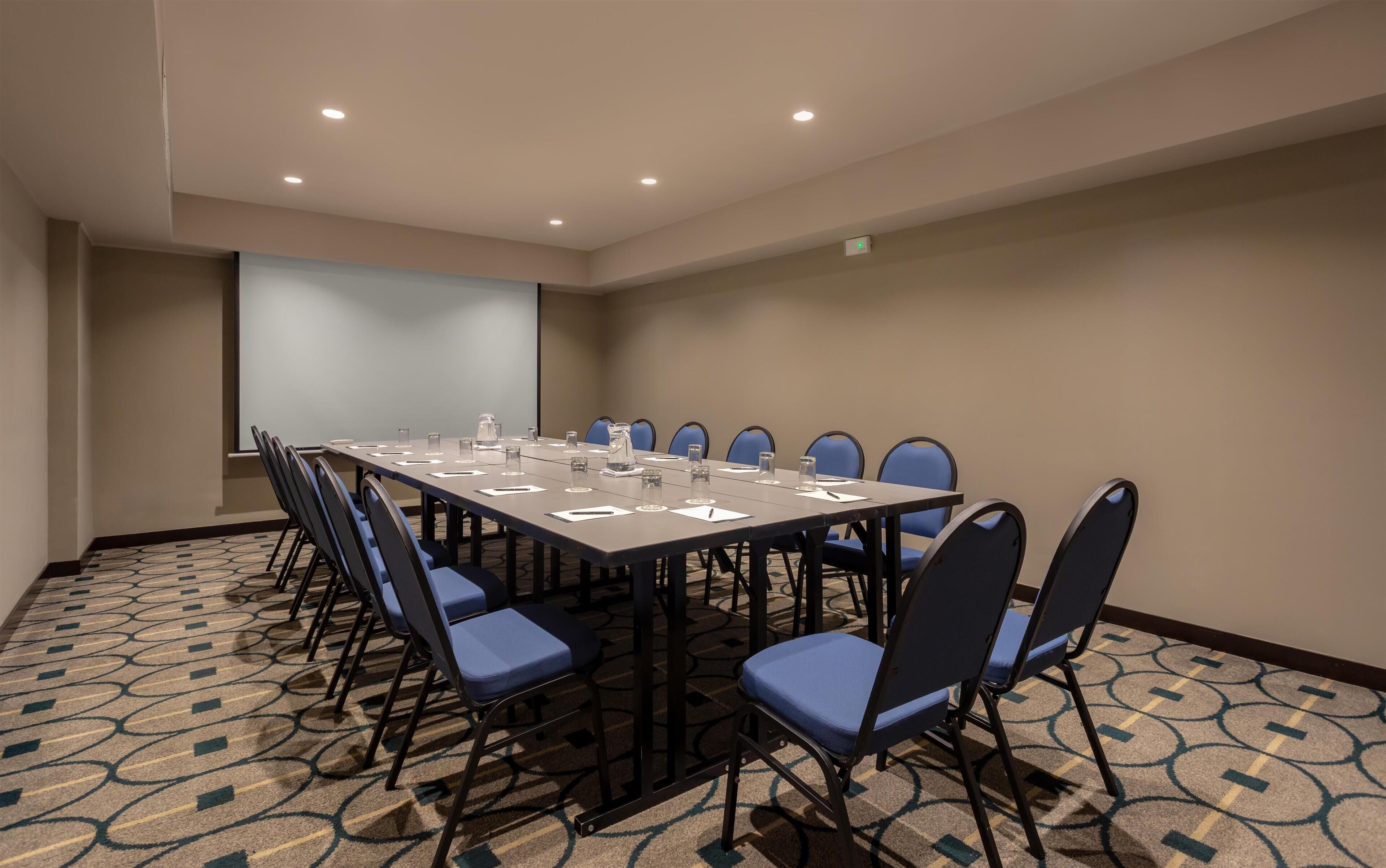 Une salle de conférence avec une longue table, des chaises bleues et un écran de projection.