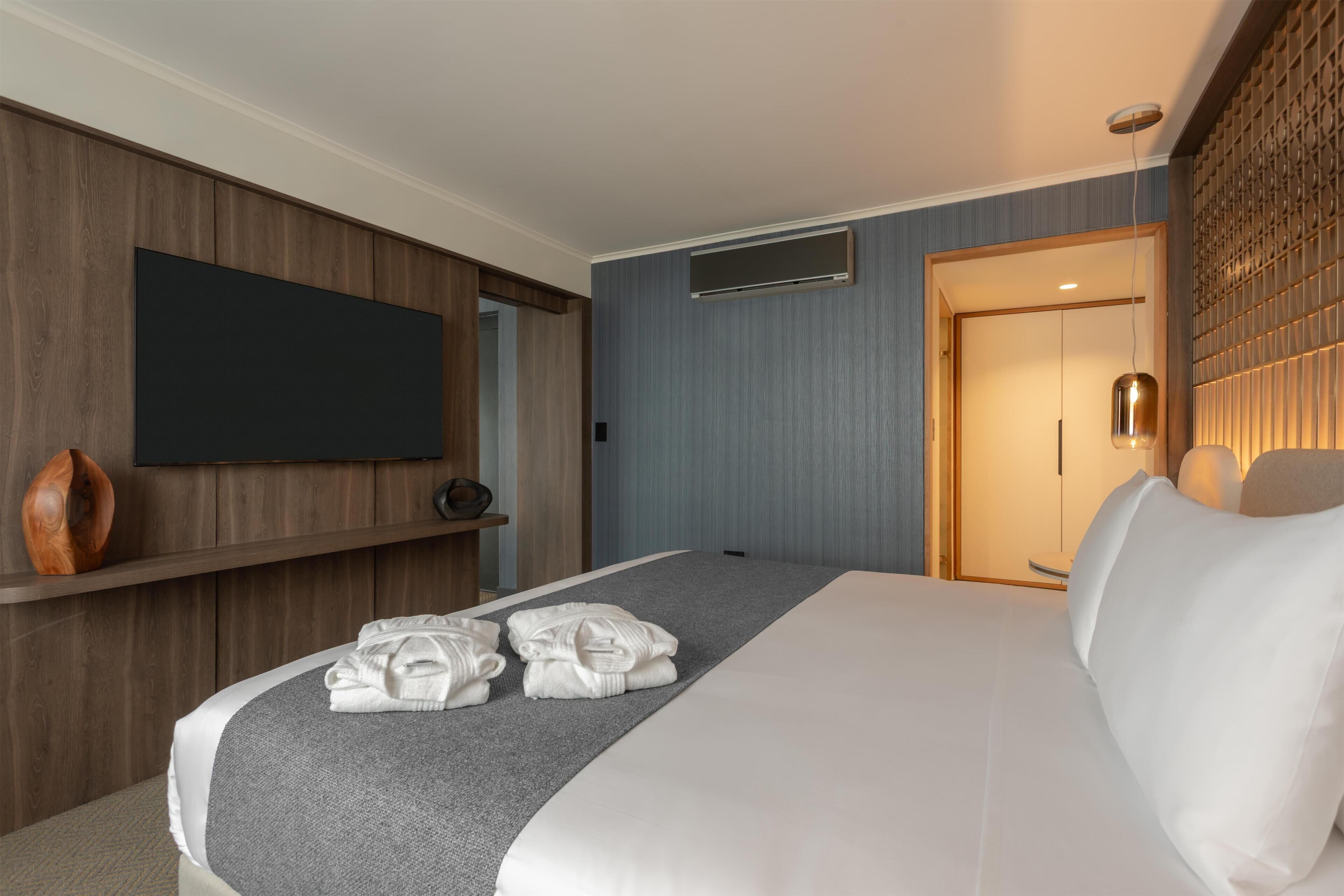 A hotel room with a large bed, a flat-screen TV mounted on the wall, a wooden panel wall, and a door leading to another room.