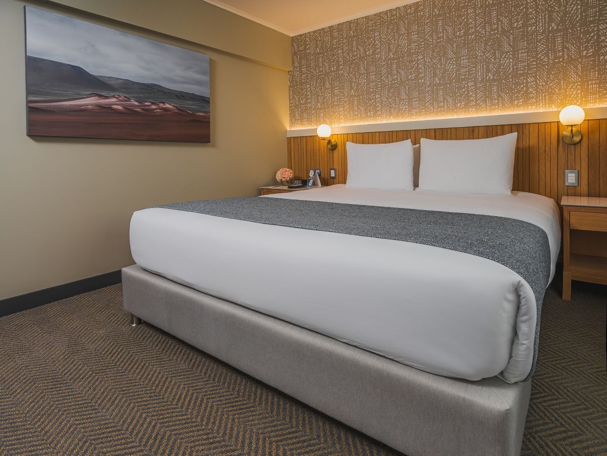 Habitación de hotel con una cama grande, cabecera con apliques de pared, mesita de noche con un teléfono y un florero, y un cuadro en la pared.