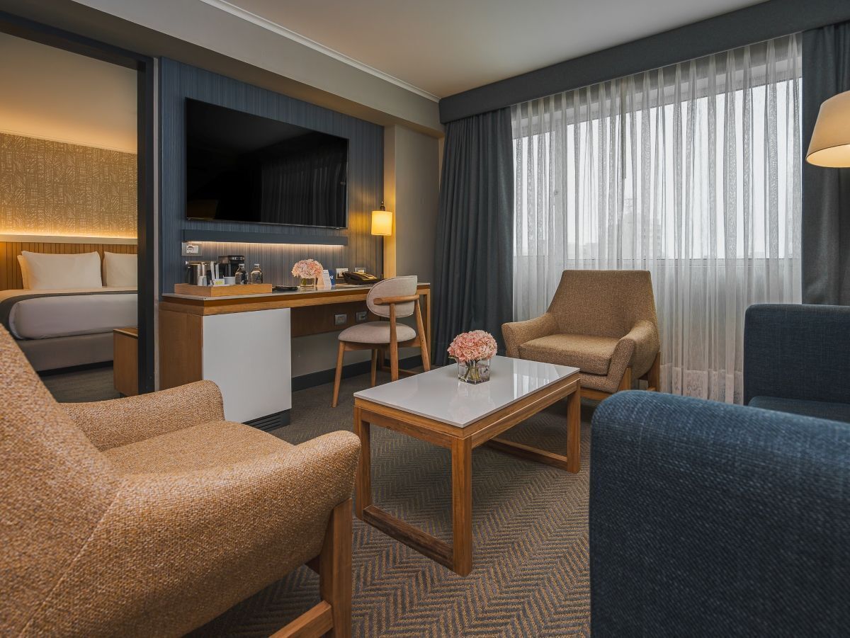 Habitación de hotel con sofá, sillón y una pequeña mesa con un florero. Hay un escritorio con una computadora y un televisor montado en la pared.