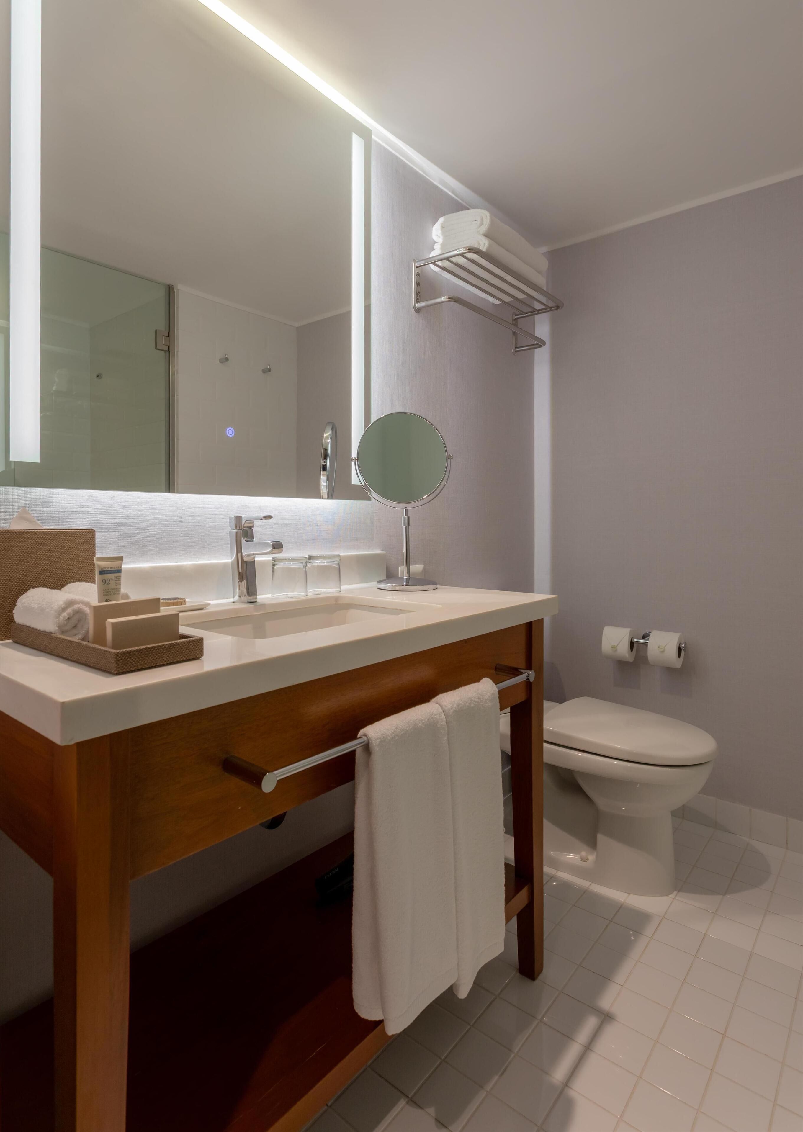 A modern bathroom with a wooden vanity, a large mirror, a round mirror, a toilet, and a towel rack.
