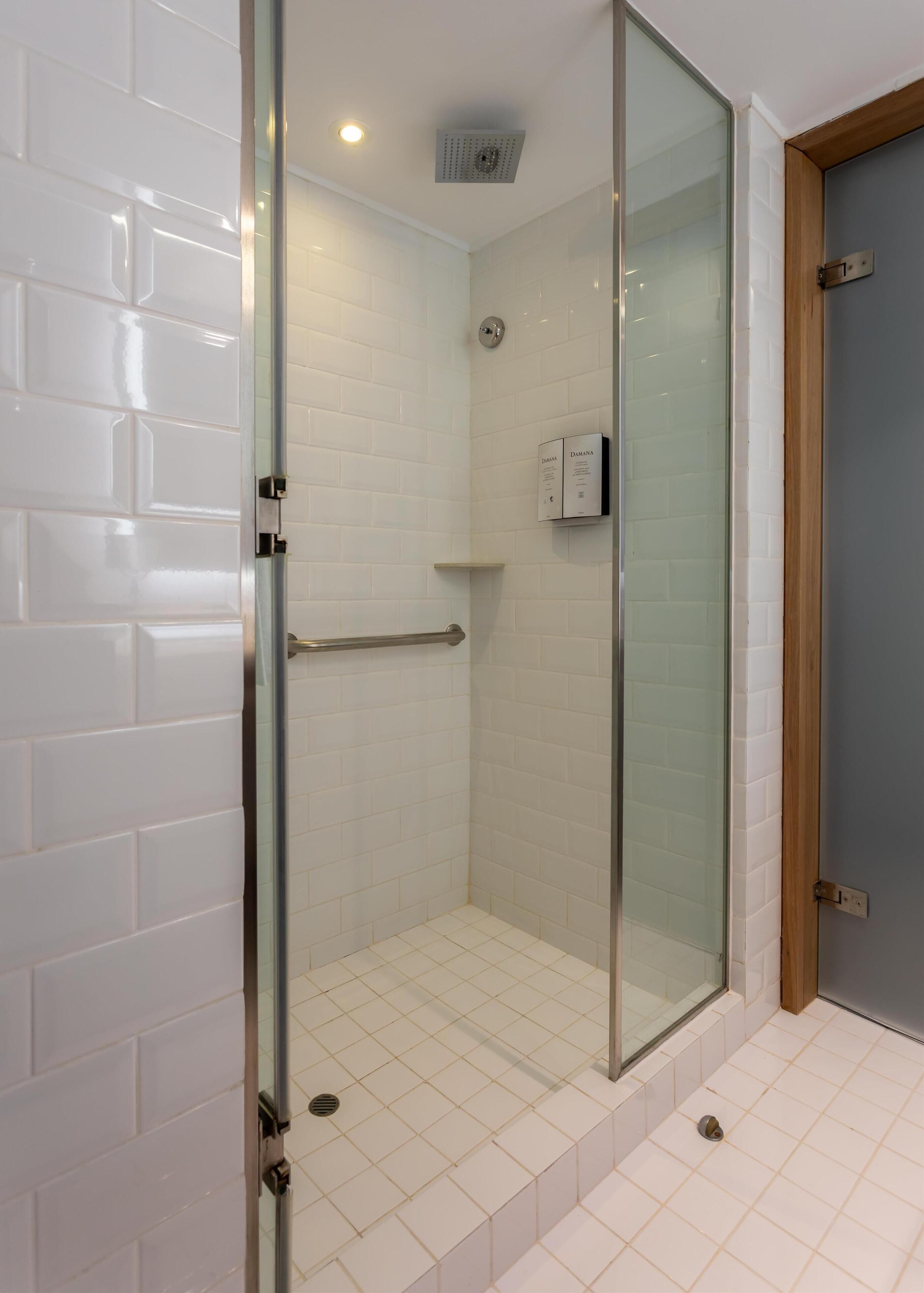 Ducha con cerramiento de vidrio, paredes y piso de azulejos blancos, baranda de metal y marco de puerta de madera.