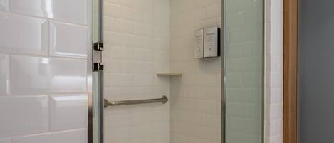 Ducha con cerramiento de vidrio, paredes y piso de azulejos blancos, baranda de metal y marco de puerta de madera.