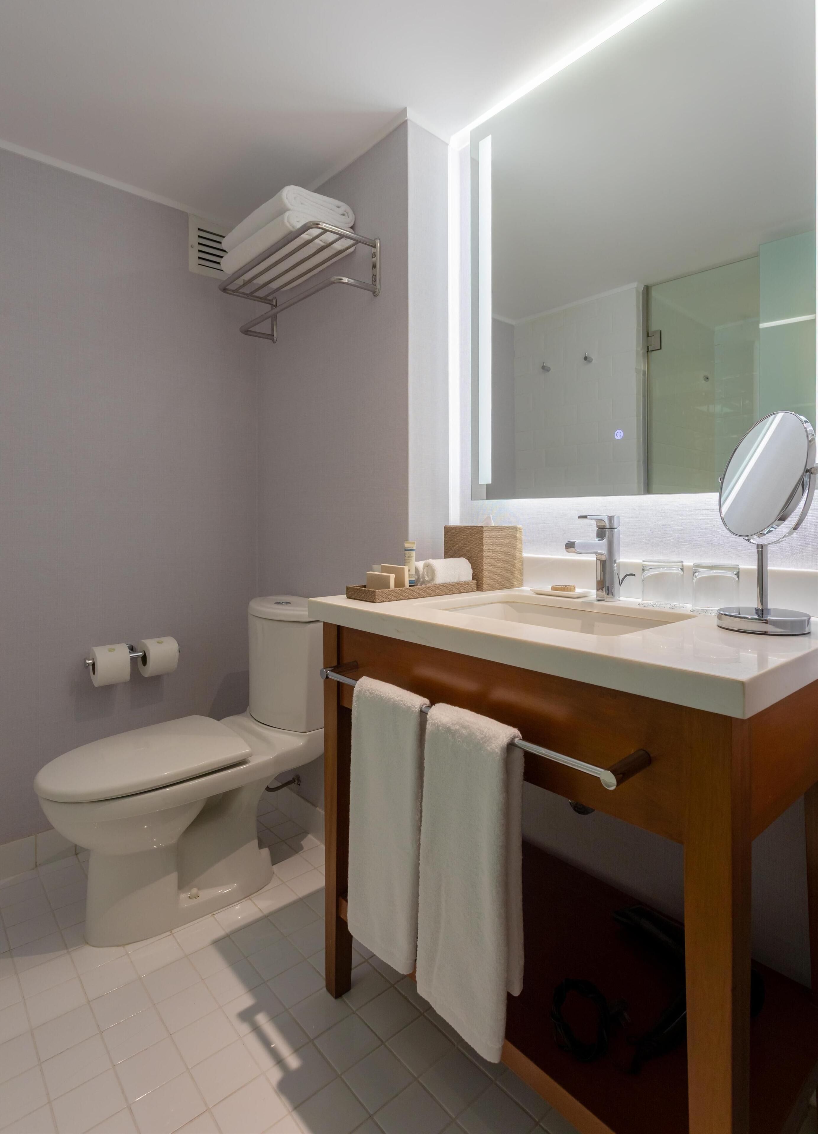 A modern bathroom with a toilet, sink, and mirror. There is a towel rack with towels, a soap dispenser, and a toilet paper holder.