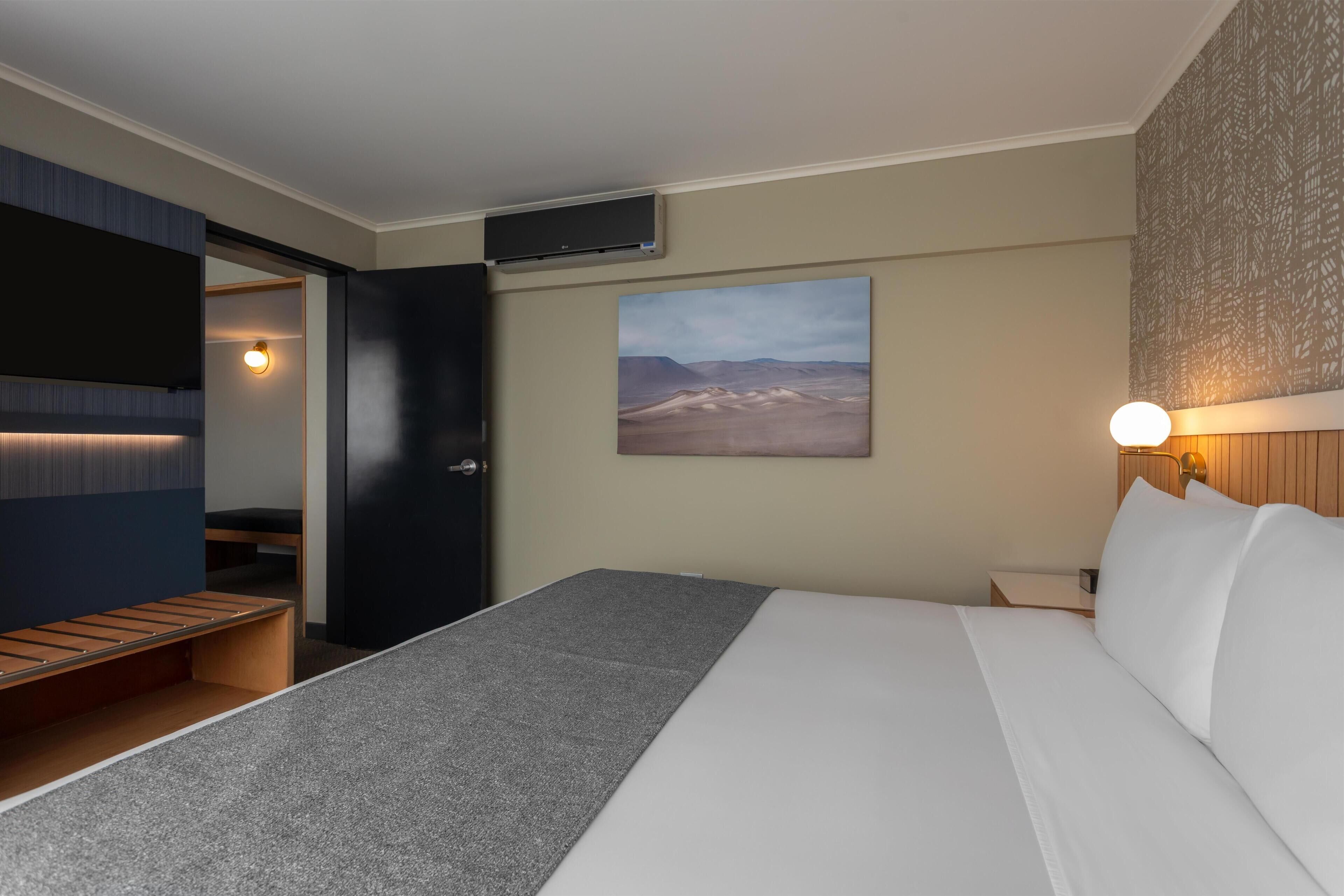 A modern hotel room with a bed, a bench, a wall-mounted TV, and a desert landscape painting.