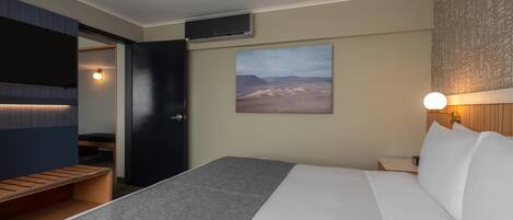 A modern hotel room with a bed, a bench, a wall-mounted TV, and a desert landscape painting.