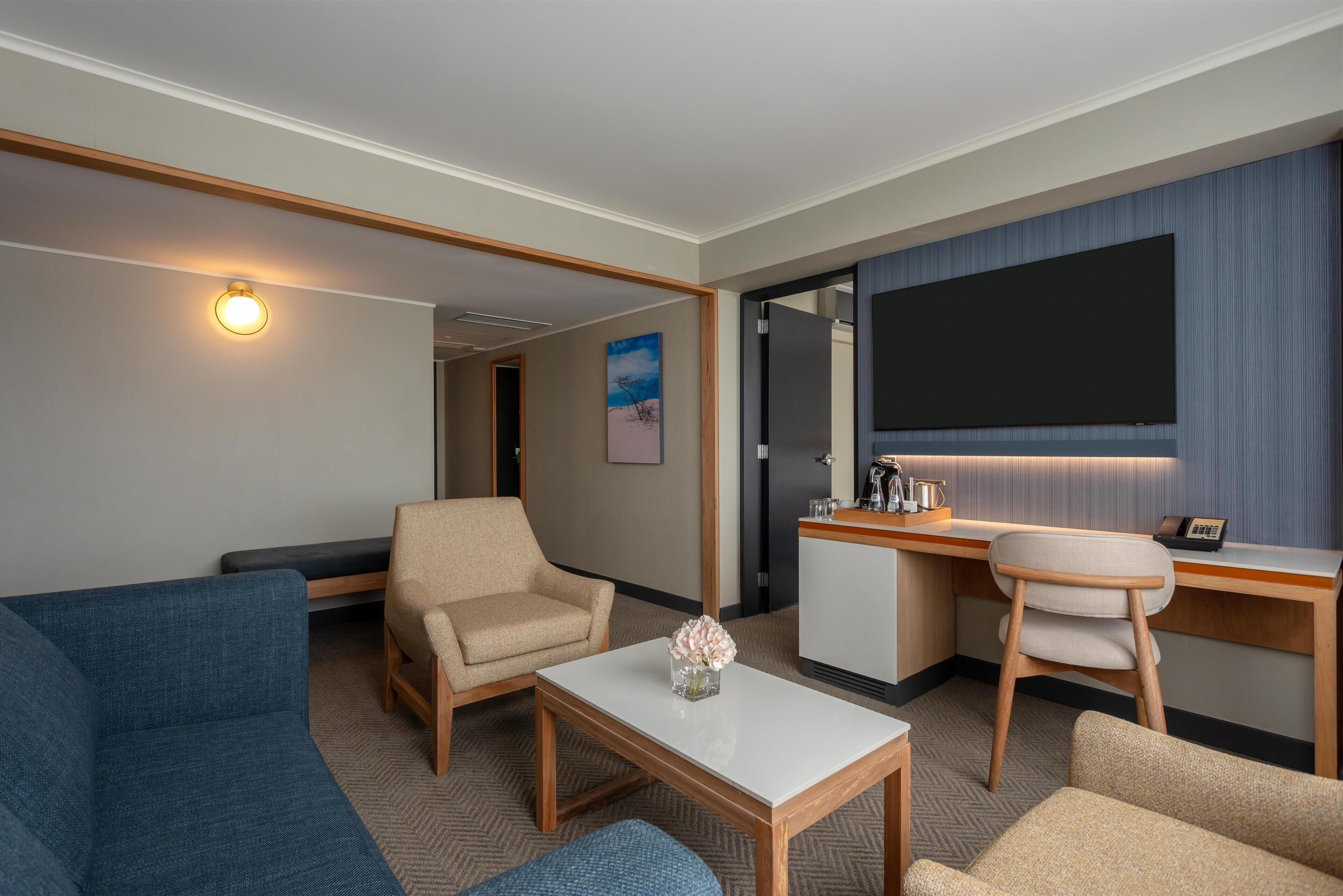 A hotel room with a sofa, armchair, desk, and a flat-screen TV.