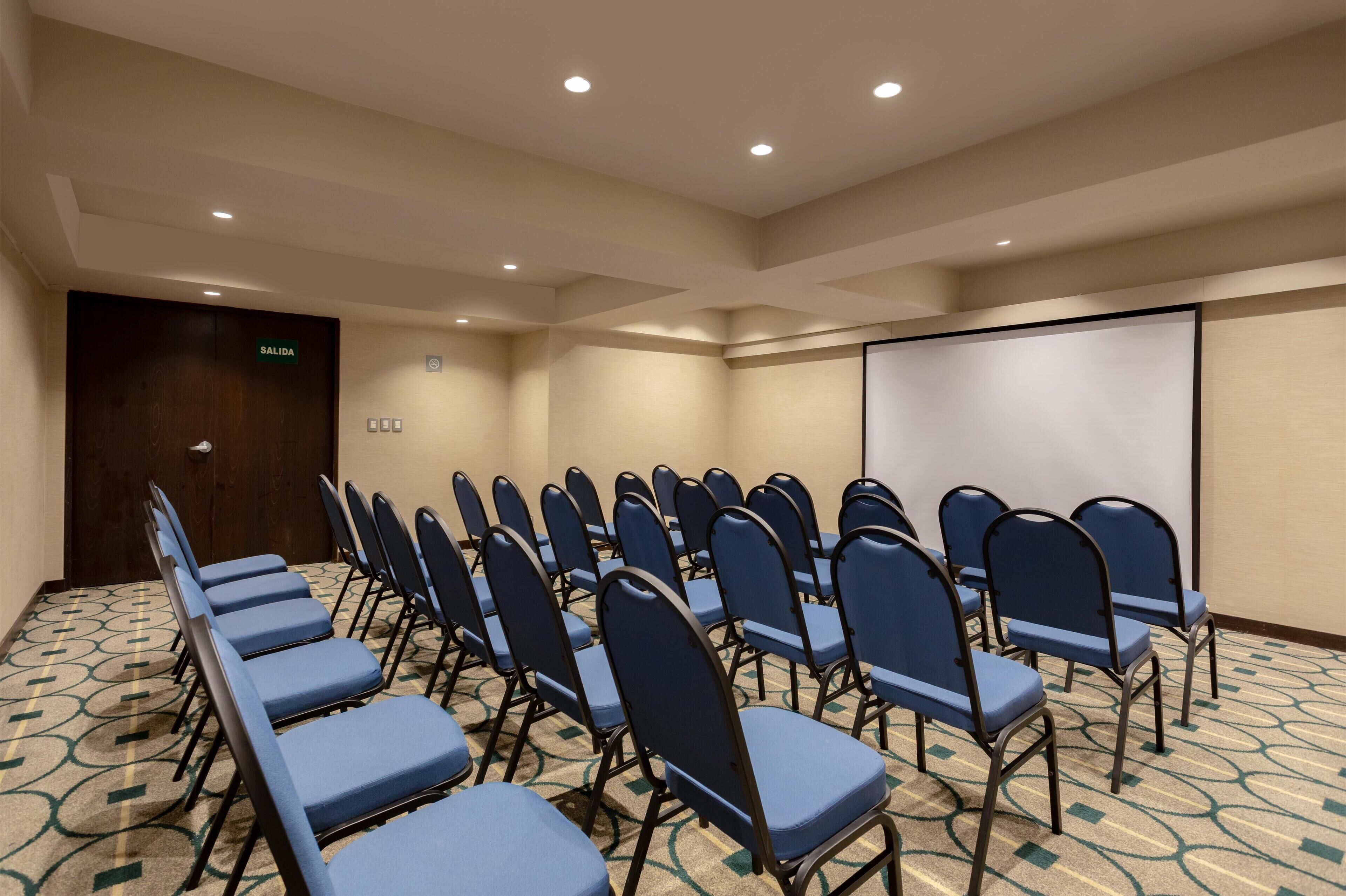 A conference room with rows of blue chairs, a projector screen, and a door labeled "SALIDA".