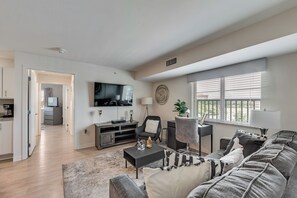 A modern living room with a flat-screen TV, a sofa, a coffee table, and a dining area with a TV and a plant. - RD402 - Home|Yard|12mi to Convention Ctr|Sleeps 6 (Richardson)