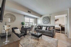 A modern living room with a grey sofa, black armchair, and a coffee table. There is a large abstract artwork on the wall and a window with blinds.