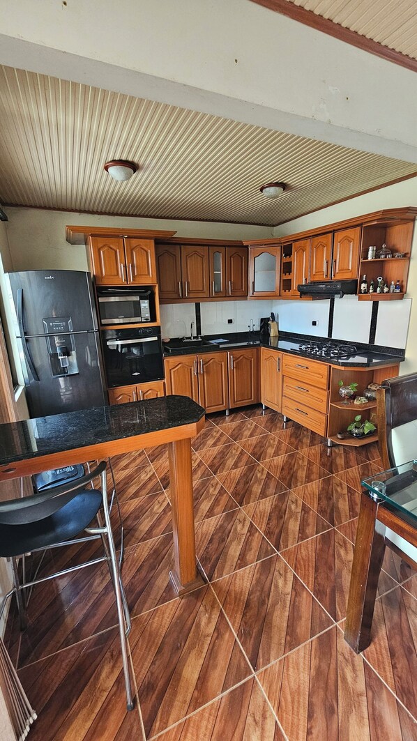 A kitchen with wooden cabinets, a refrigerator, stove, and a dining area with a chair.