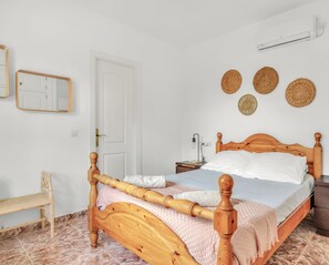 A neatly arranged bedroom with a wooden bed, a bedside table, a nightstand, a chair, and a wall-mounted air conditioner.