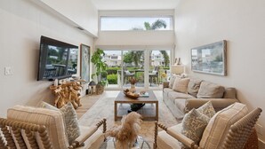 A spacious living room with a large flat-screen TV, comfortable seating, a coffee table, and a balcony with a view of palm trees and buildings. - Coastal Oasis, Lovely 3-bedroom condo with AC in Holmes Beach (FL - Holmes Beach)