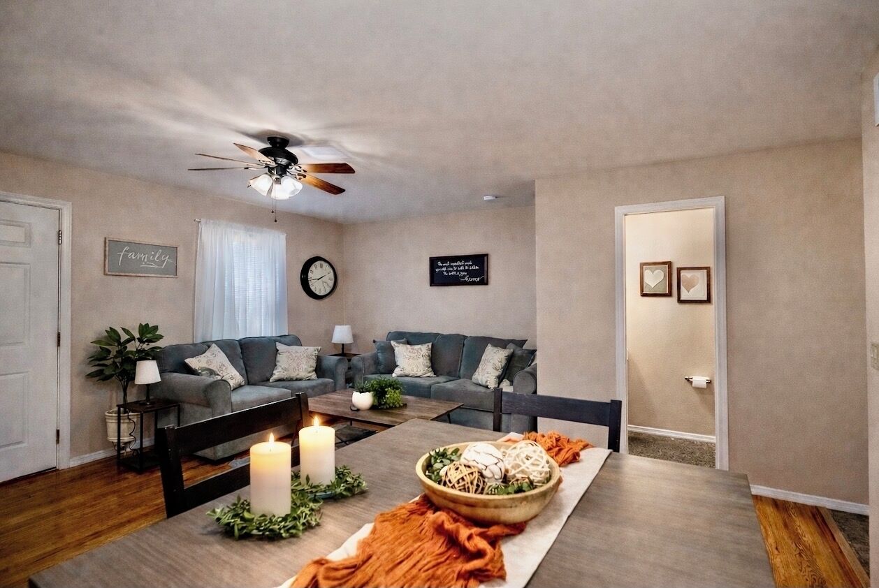 Un salon avec une table à manger dressée pour le repas, un ventilateur de plafond, un canapé et une horloge murale.