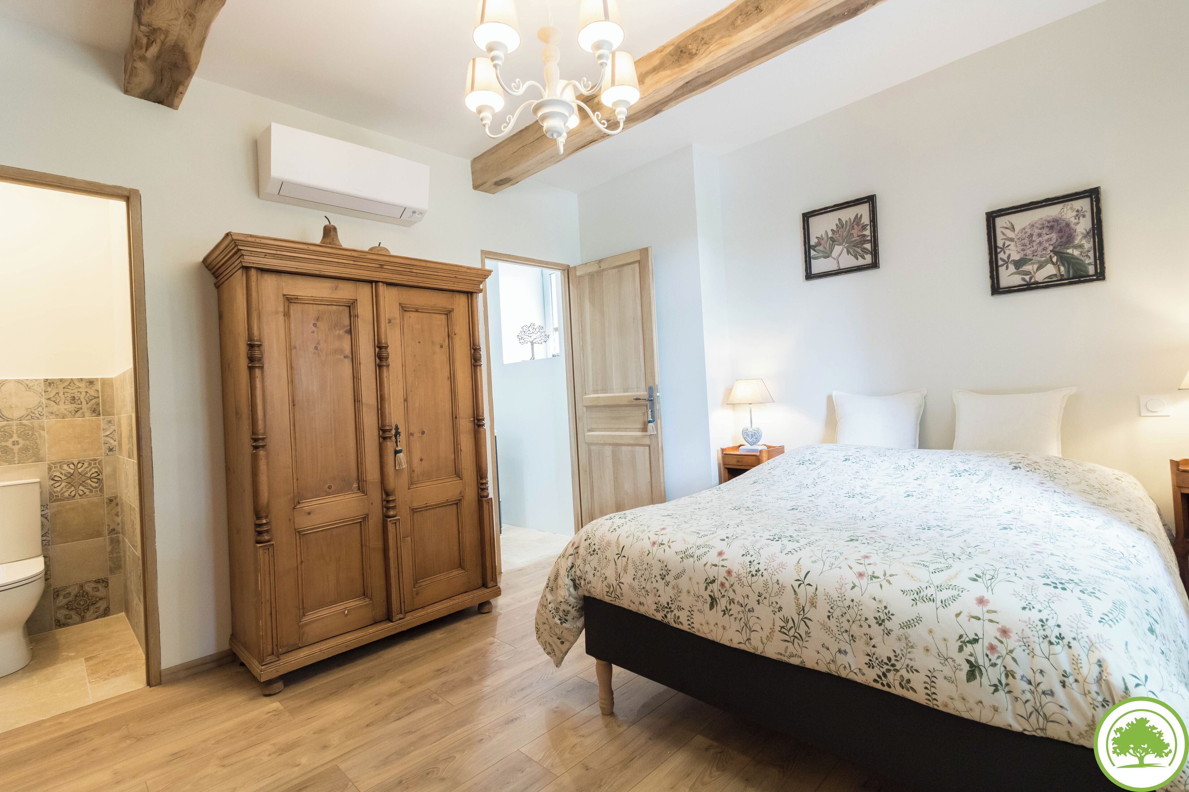 A bedroom with a wooden wardrobe, a bed with floral bedding, a chandelier, and two framed botanical prints on the wall.